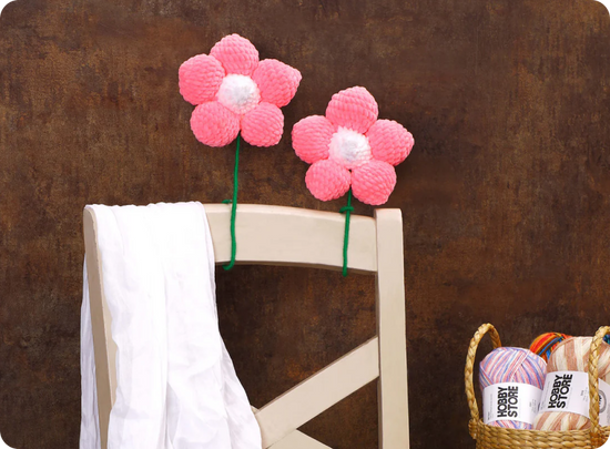 Two pink crocheted flowers on a chair with a basket of yarn in the background.